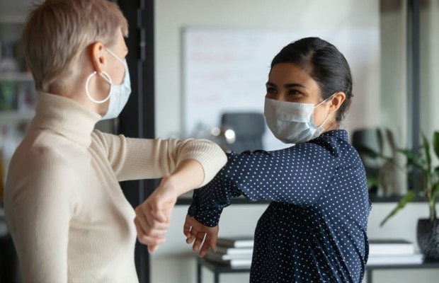 Lächelnde weibliche Kollegen, die Schutzmasken tragen Grußmasken und Ellbogen am Arbeitsplatz springen, Frauen-Mitarbeiter in Gesichtsbeuteln schützen vor COVID-19 Coronavirus im Amt, Gesundheitskonzept